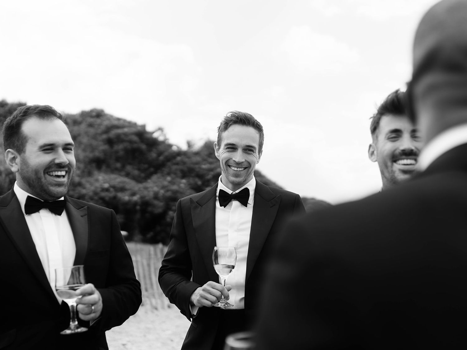Men in tuxedos share laughs and drinks at an outdoor celebration.