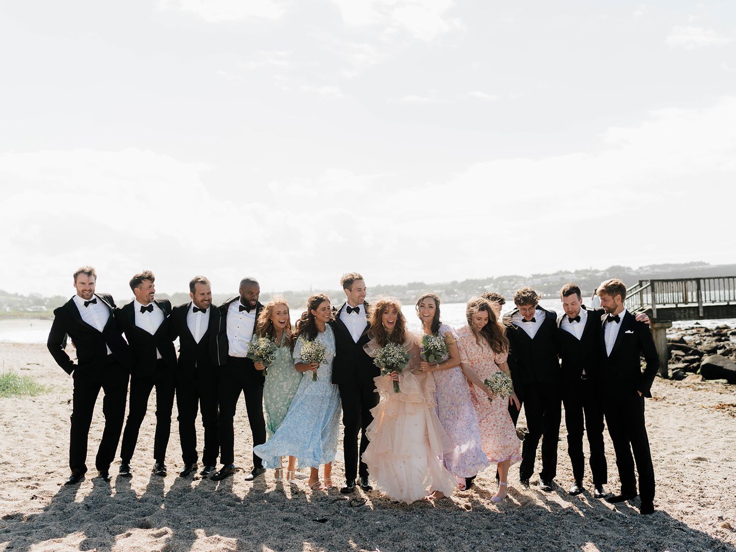 Wedding party in formal attire lined up on beach with groomsmen in black and bridesmaids in pastel dresses.