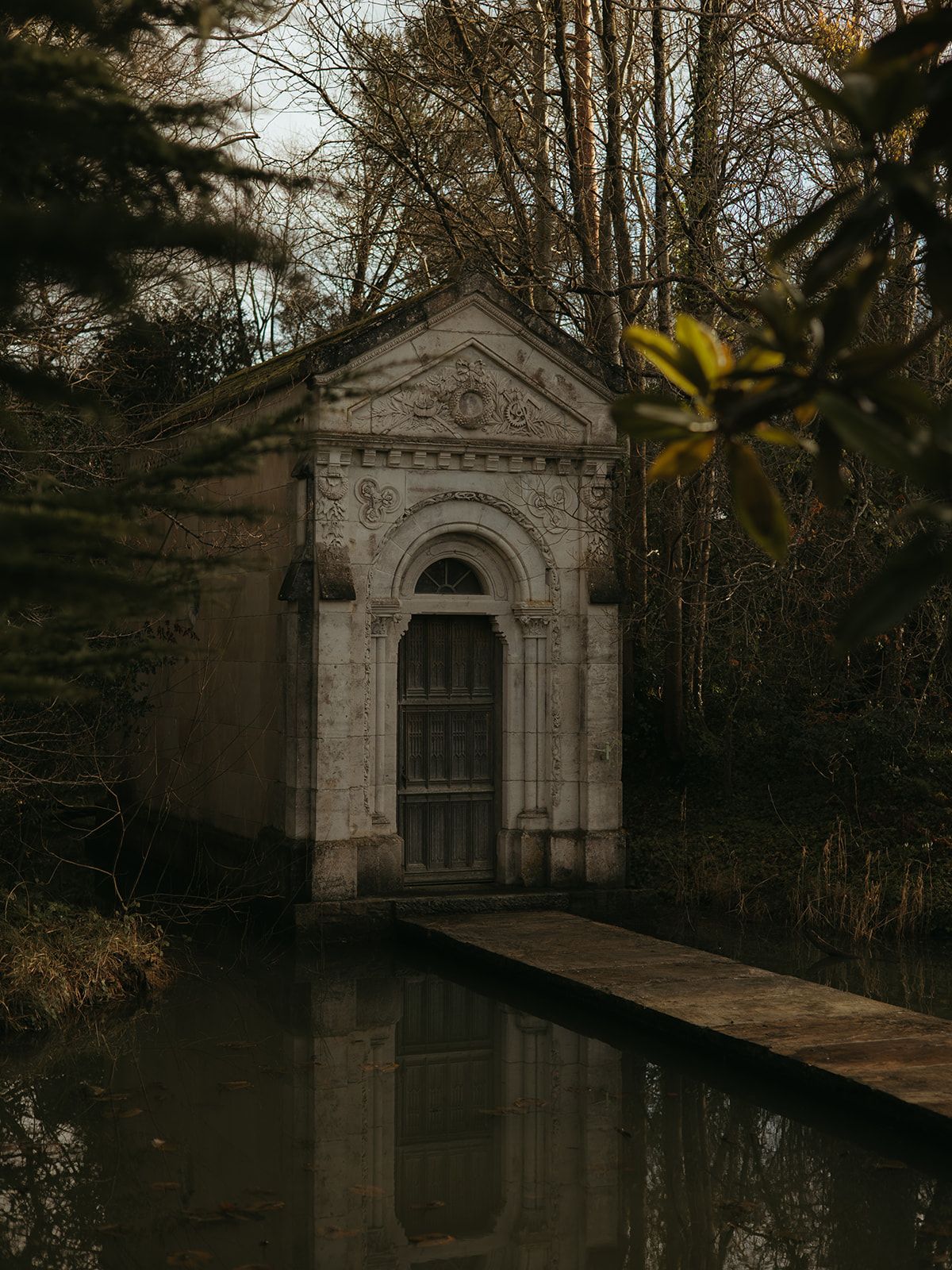 A small stone mausoleum with ornate architecture reflects in still waters surrounded by bare trees.