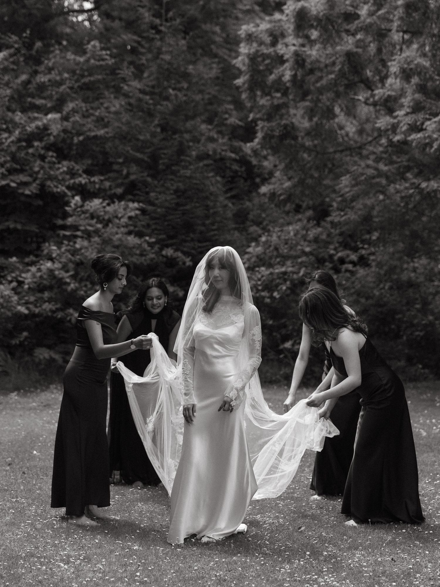 Black and white photo of a bride in a white gown and veil surrounded by bridesmaids in black dresses outdoors.