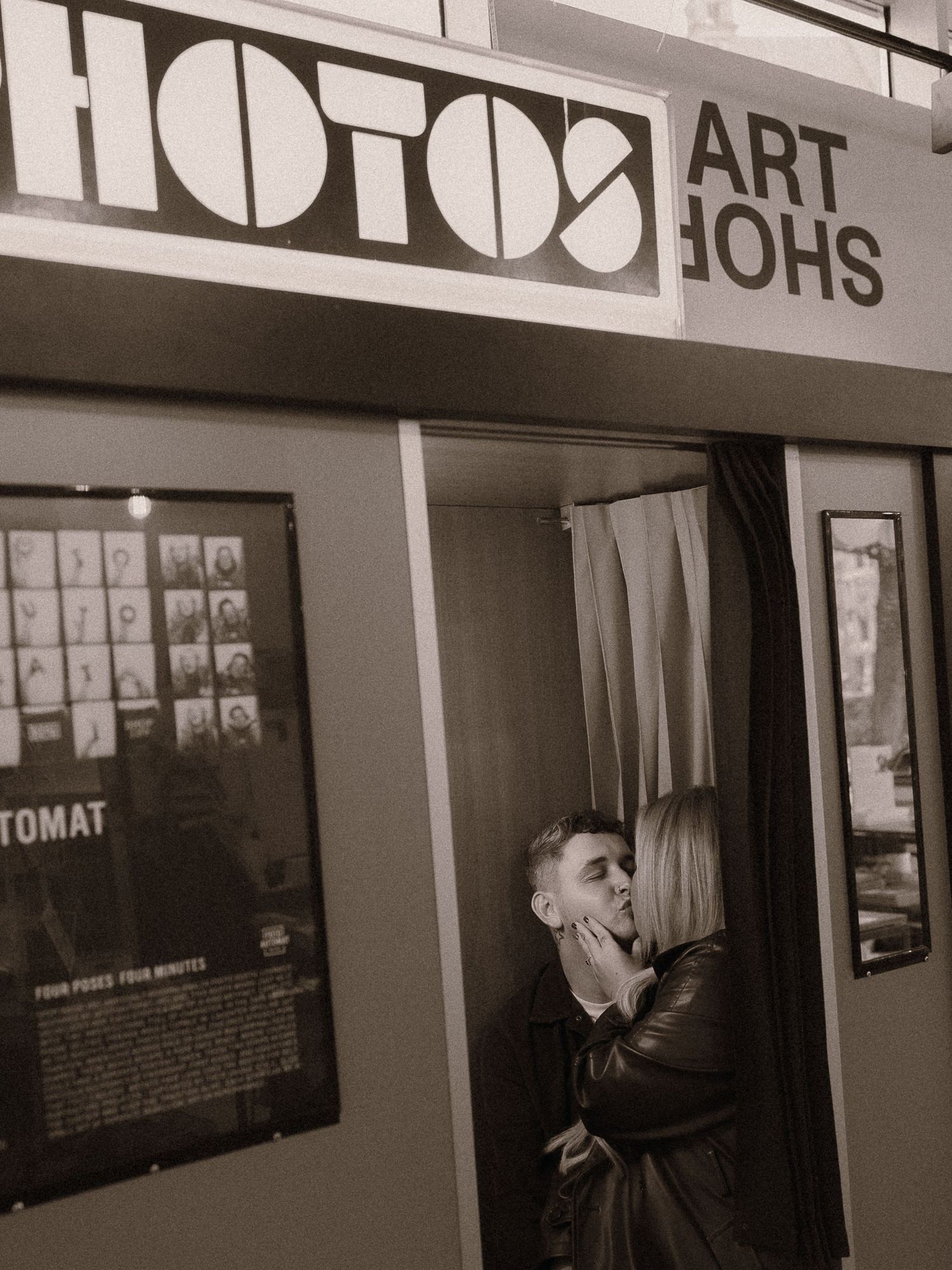 Couple sharing a romantic kiss outside a vintage photo booth art show in a sepia-toned street photography shot.