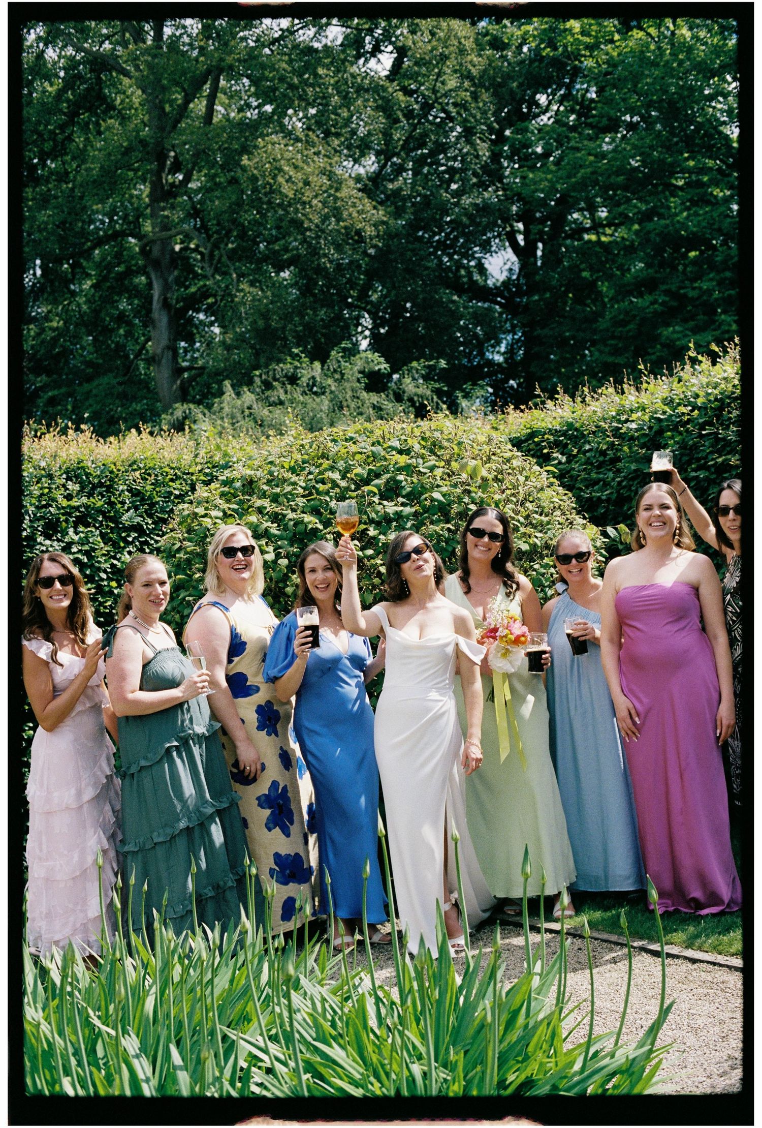 Bride in white dress posing with seven bridesmaids in colorful gowns in a garden setting.