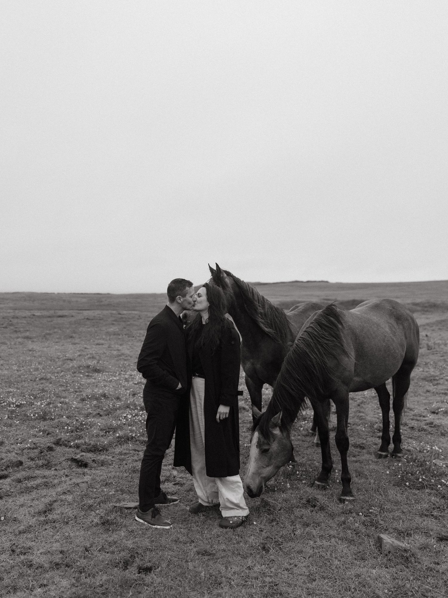 A couple shares a kiss while standing beside two horses in an open field, captured in black and white.
