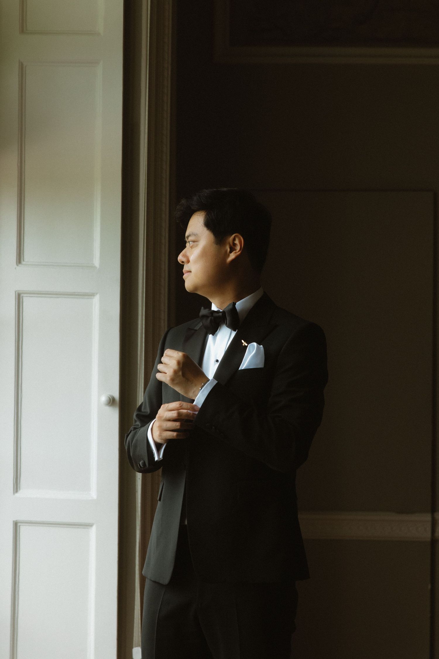 Man in black tuxedo and bowtie gazes contemplatively through window in elegant formal setting.