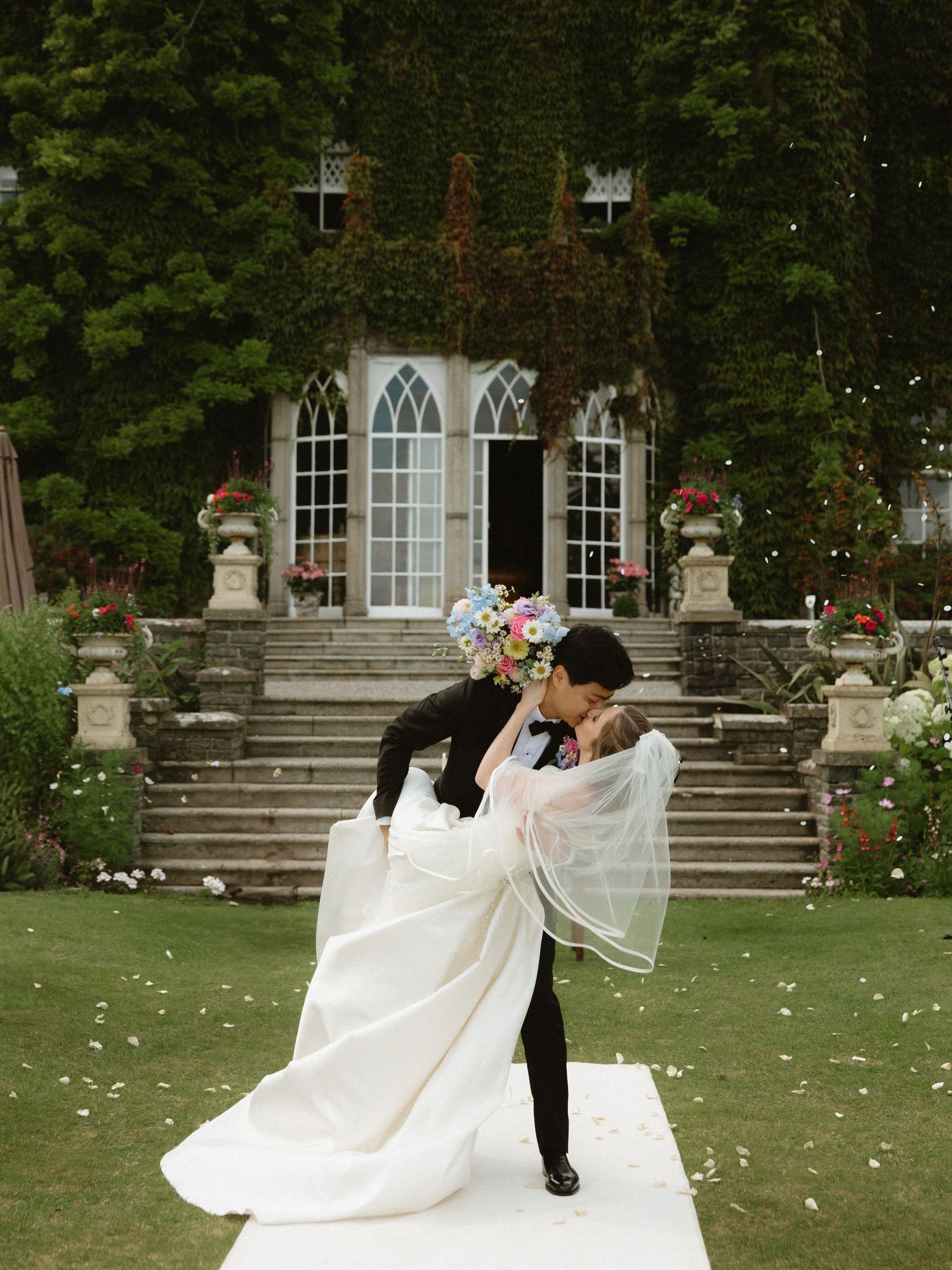 Romantic wedding couple shares a dip kiss on stone steps of a garden venue with rose petals scattered around them.