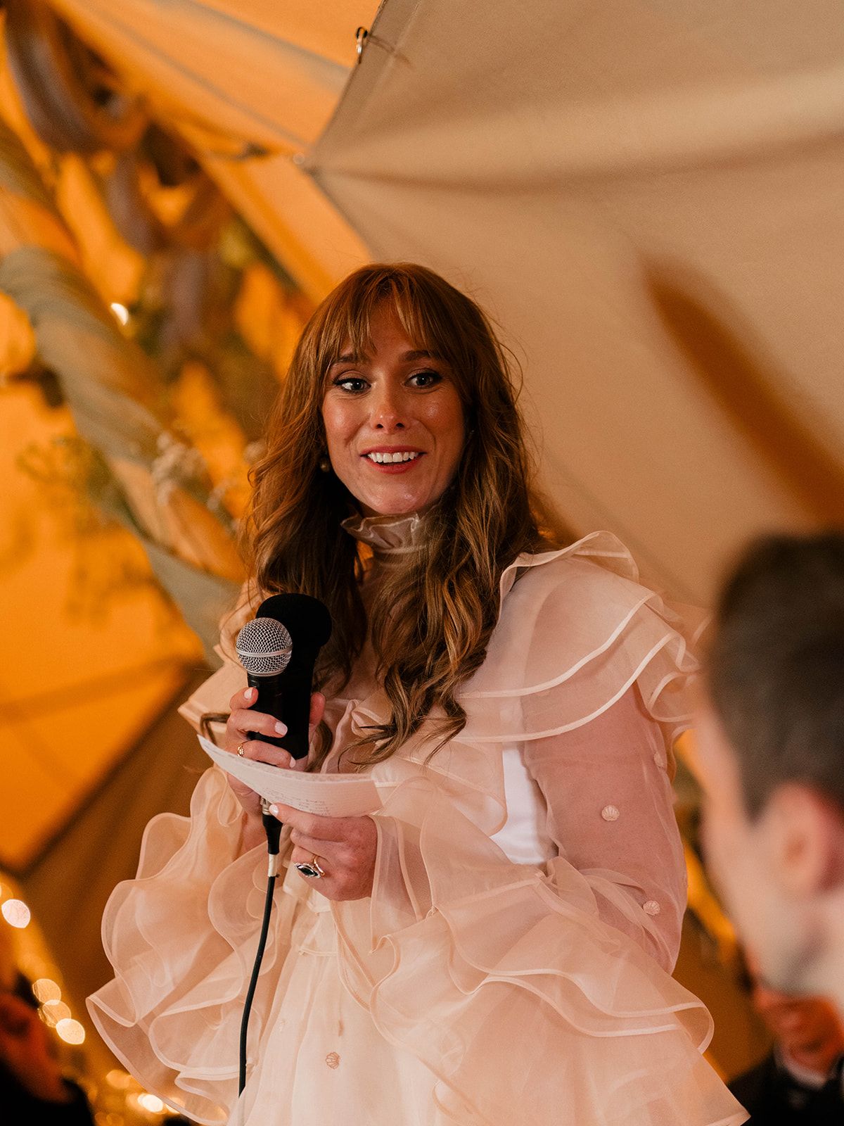 Bride in ruffled white dress holds microphone while giving speech at reception.