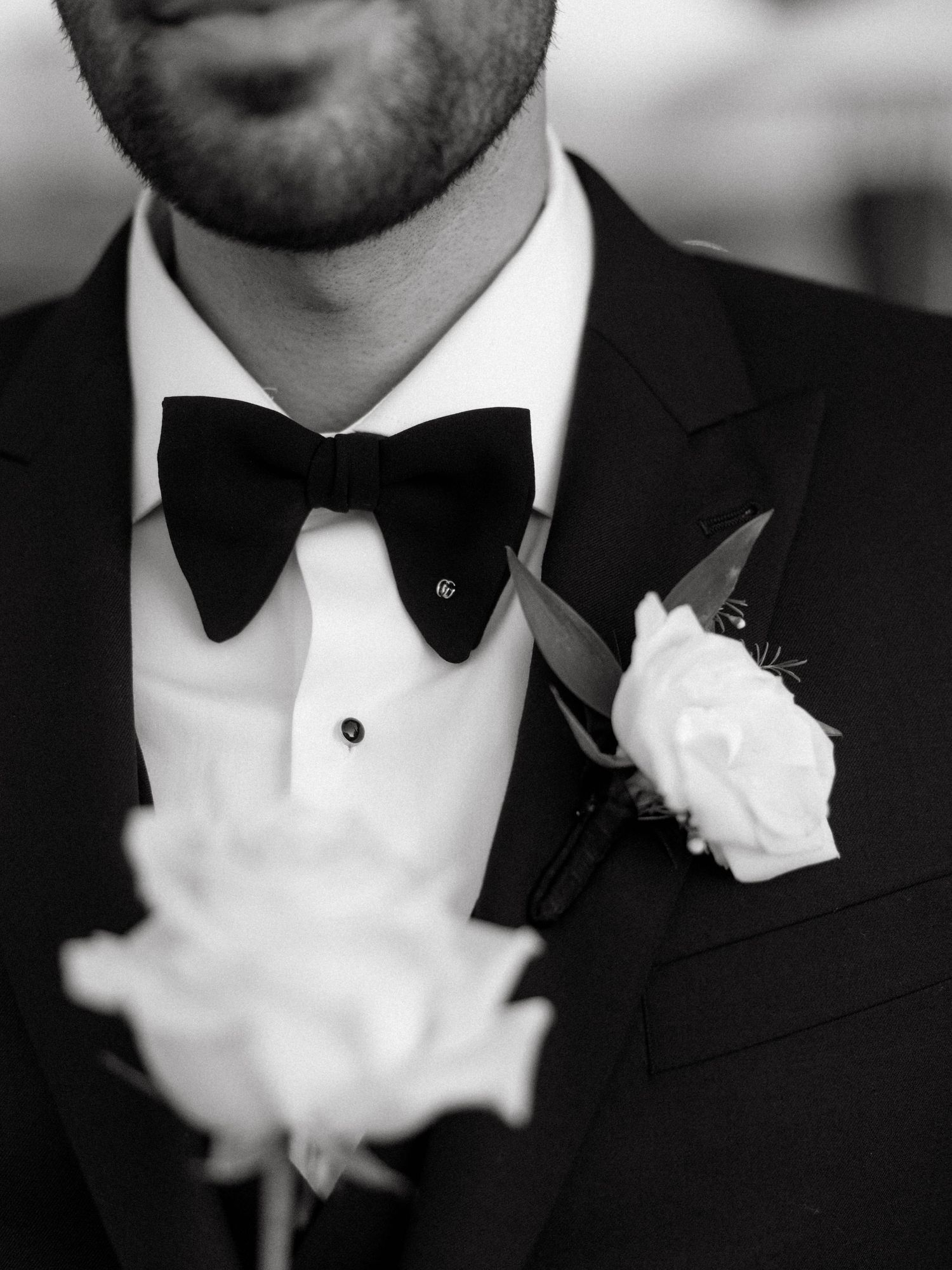 Black and white image of a formal tuxedo with black bow tie and white rose boutonniere.