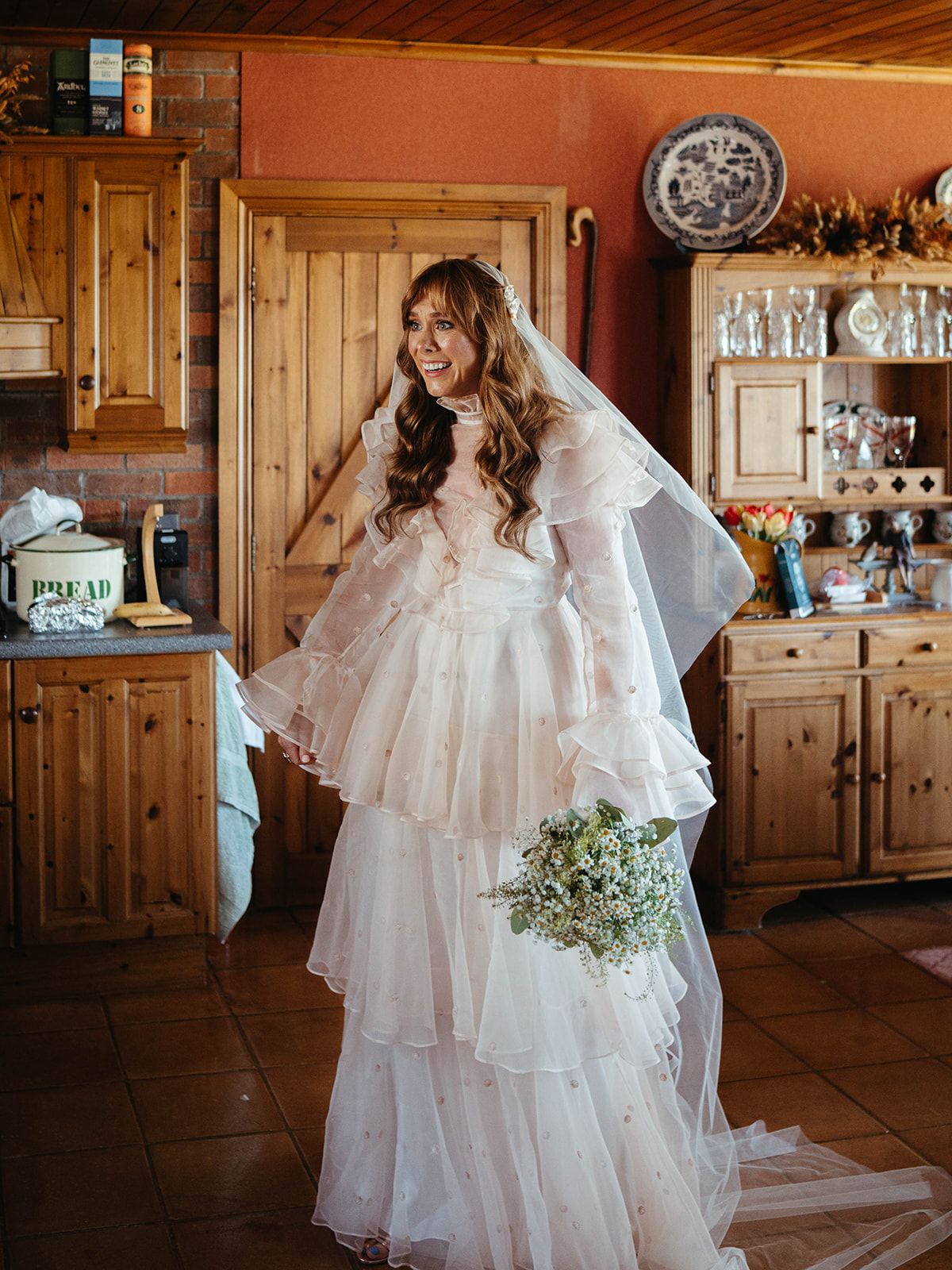 A bride in a layered white dress smiles joyfully in a rustic kitchen setting.