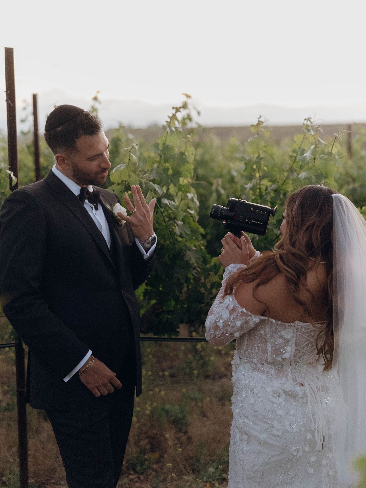 A wedding couple shares a fun moment at a vineyard as the bride turns her camera toward the groom.