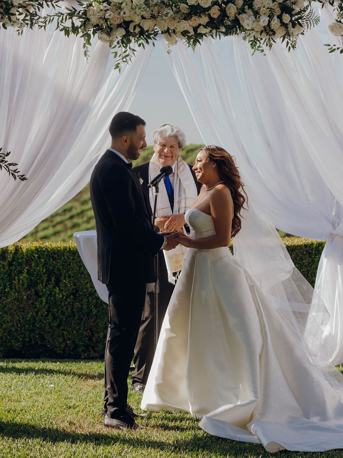 Beautiful outdoor wedding ceremony under a floral arch with flowing white drapes on a sunny day.
