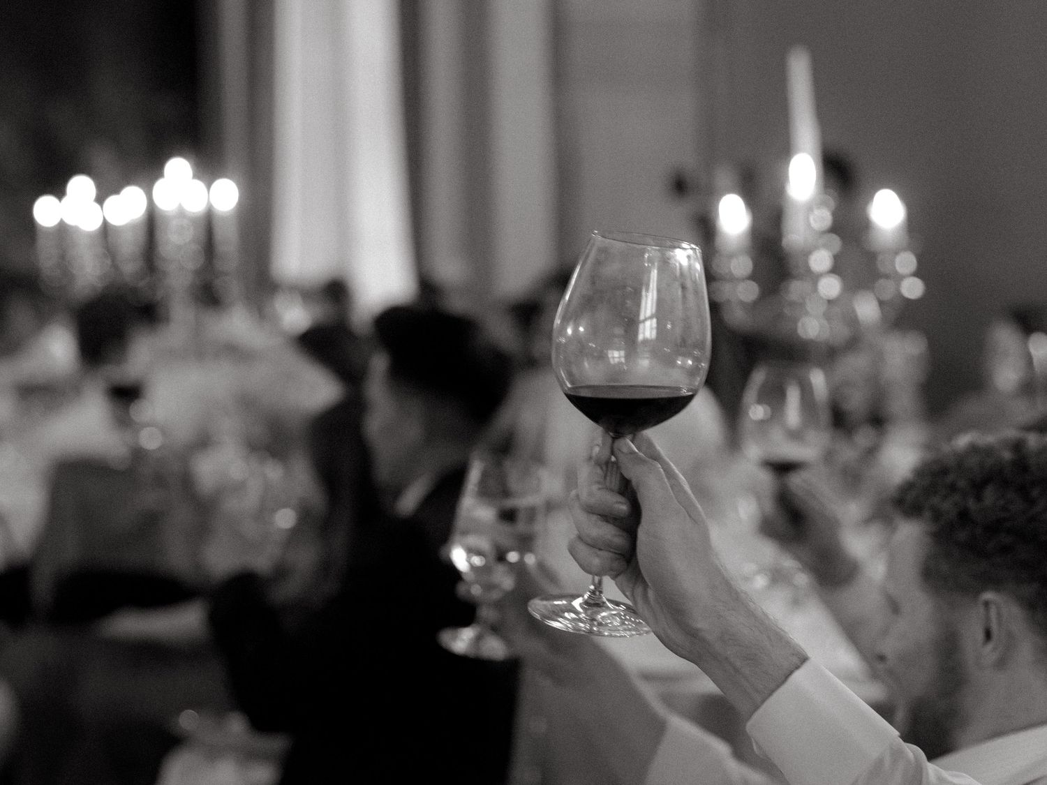 Hand holding up a glass of red wine during a toast at an elegant dinner event.