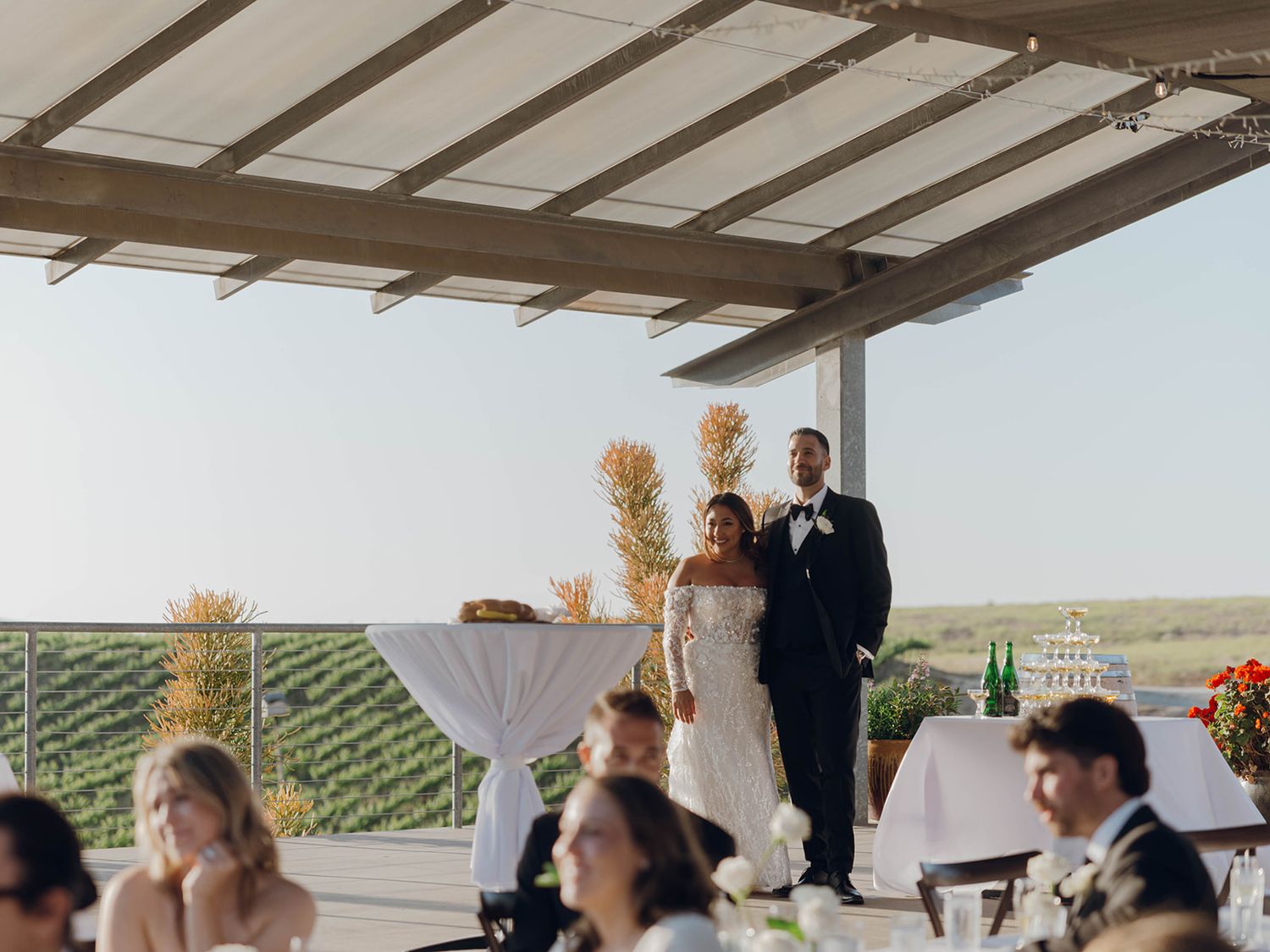 An outdoor wedding reception at a vineyard with guests seated at tables under a covered patio with scenic views.