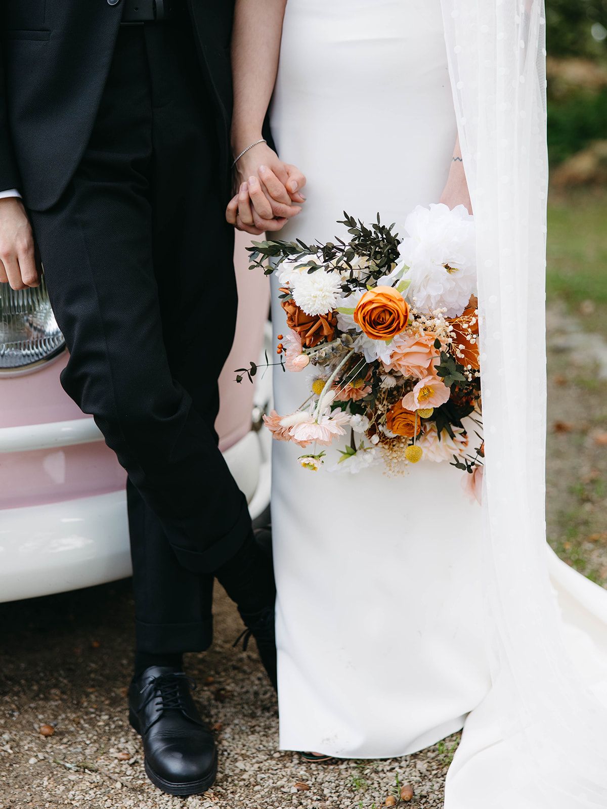 A wedding bouquet with orange roses and white flowers held against a flowing white dress next to formal black shoes.