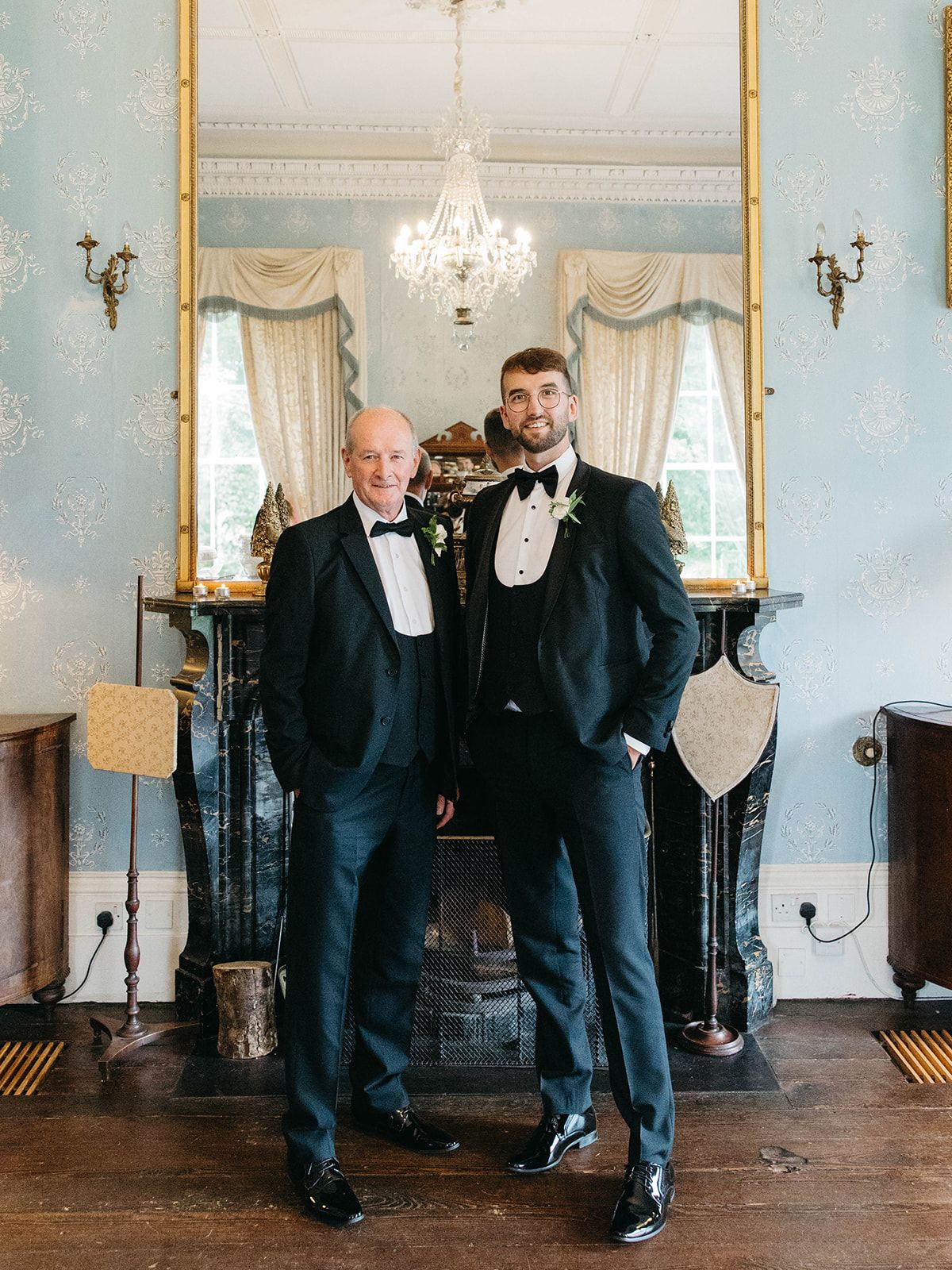 Two people in black tuxedos stand in an elegant room with blue walls, ornate fireplace and crystal chandelier.