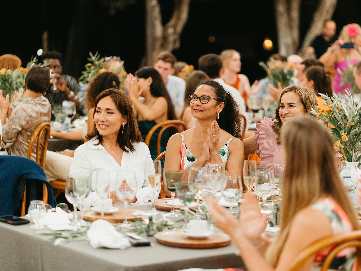 Guests enjoy an elegant outdoor dinner party with wine glasses and floral centerpieces on long tables at dusk.