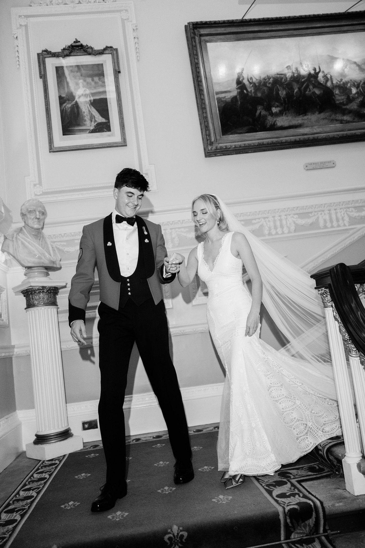 A couple in formal wedding attire dance and laugh together in an elegant ballroom with framed artwork on the walls.