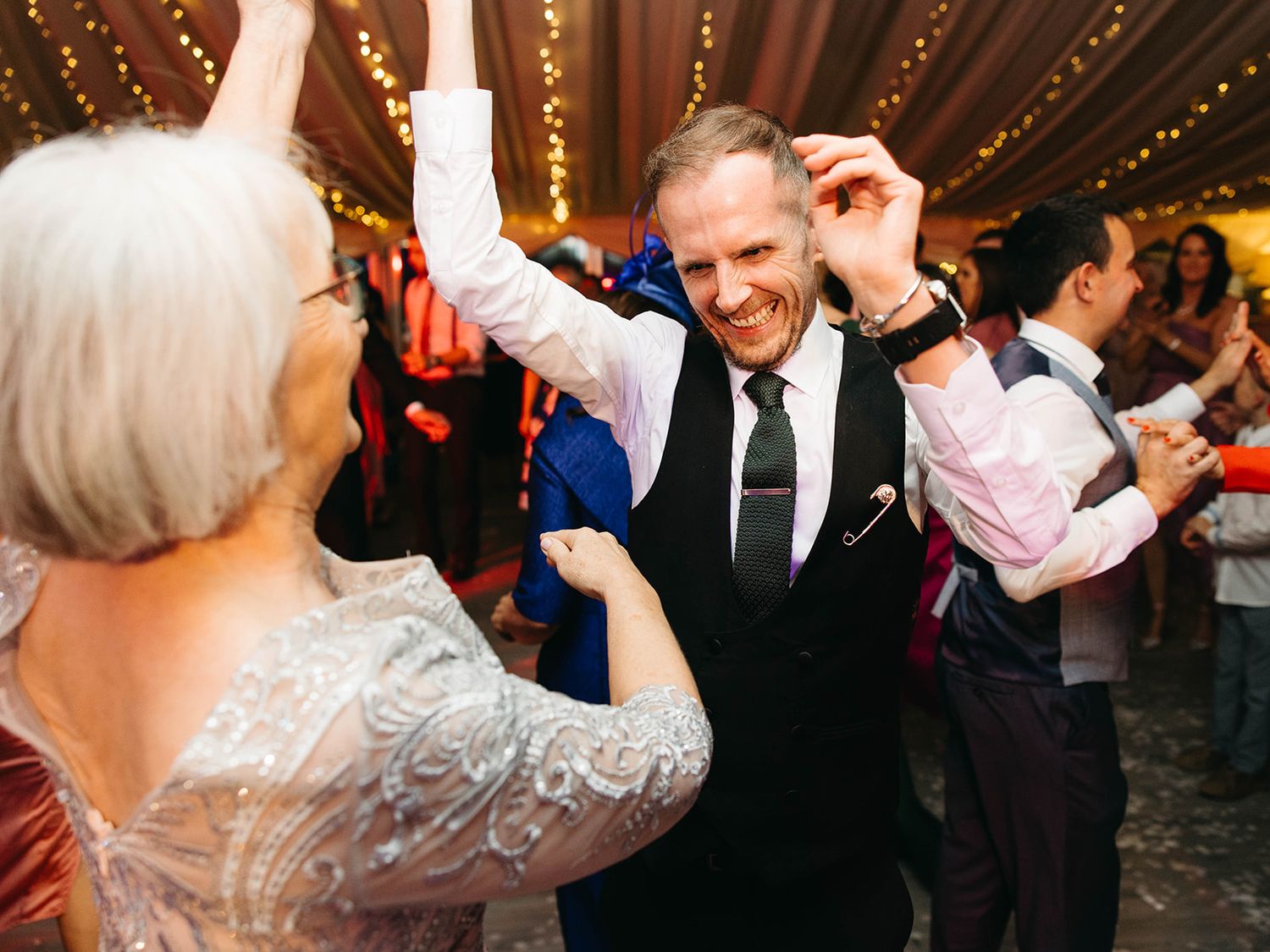 Dancing partygoers celebrate under warm string lights at an indoor reception venue.