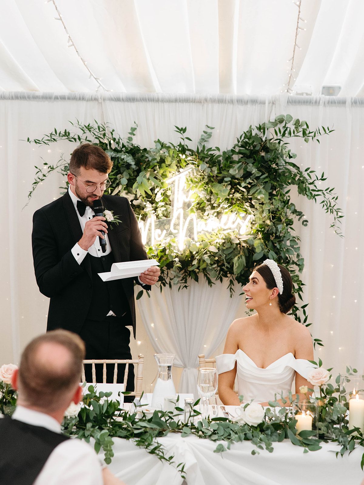 A formal wedding reception speech delivered at a decorated head table with greenery and candlelight in a white tent setting.