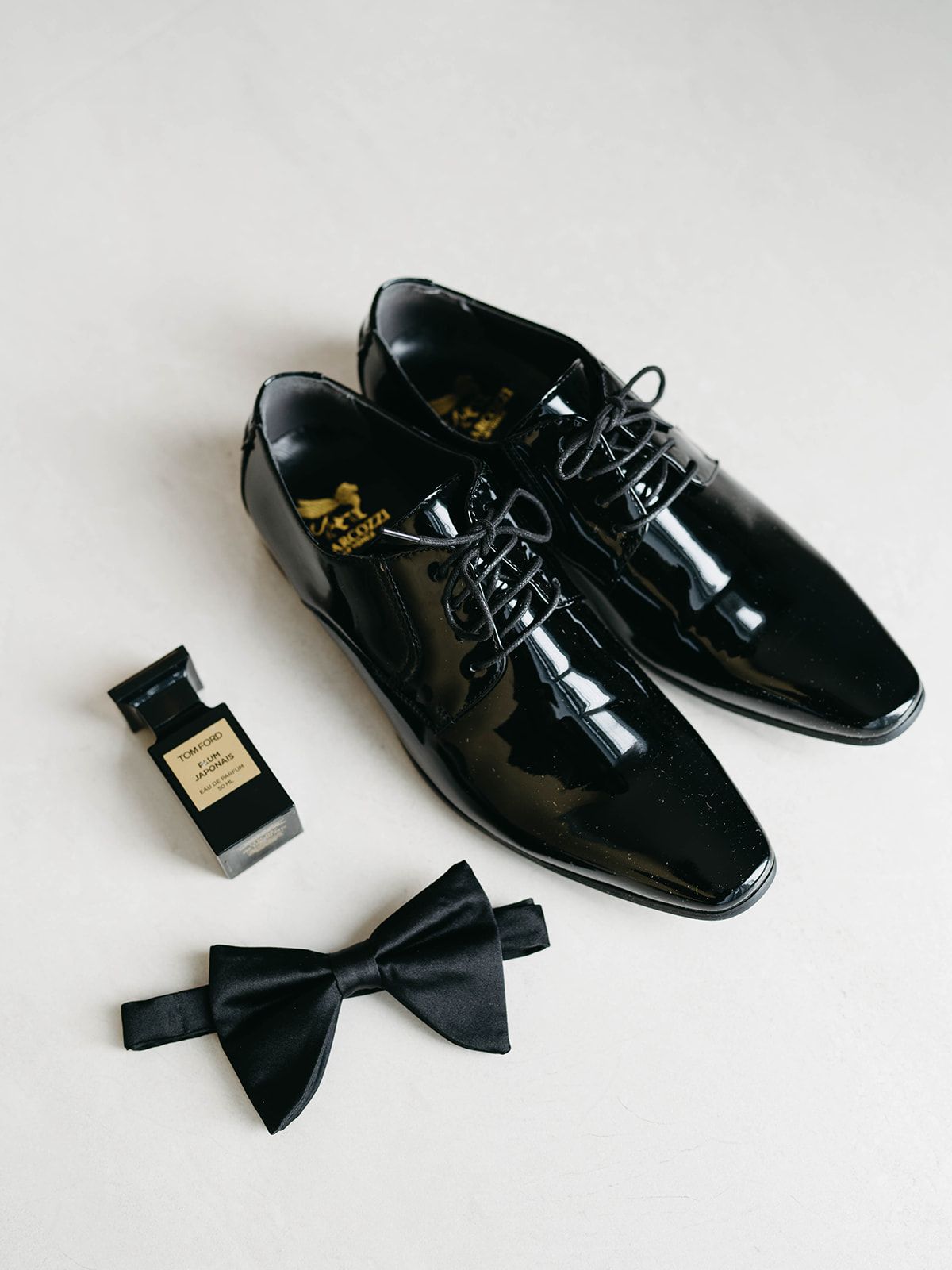 Black patent leather dress shoes with matching black bow tie and cologne bottle on white background.