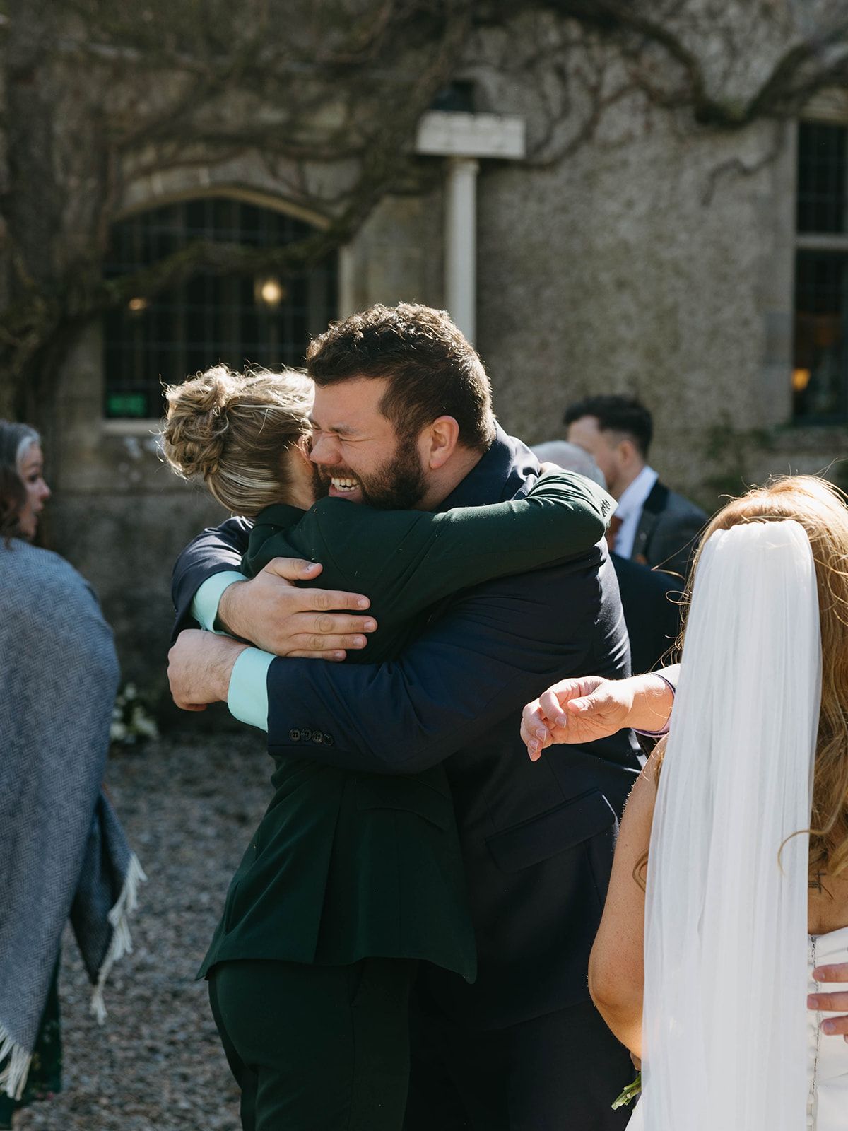 Two wedding guests share a warm embrace during an outdoor celebration at a stone venue.