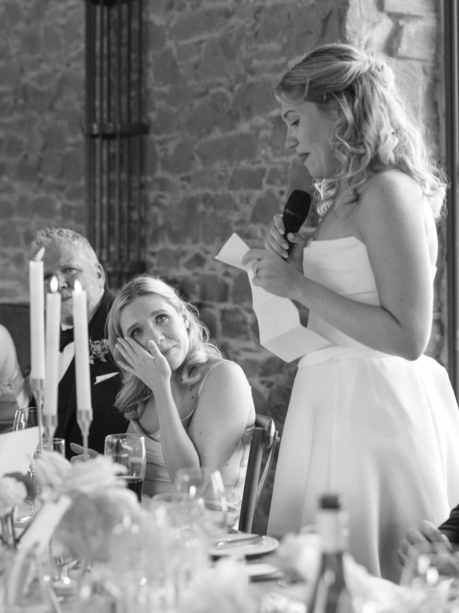 Wedding guests share emotional reactions during speeches at a stone-walled reception venue.