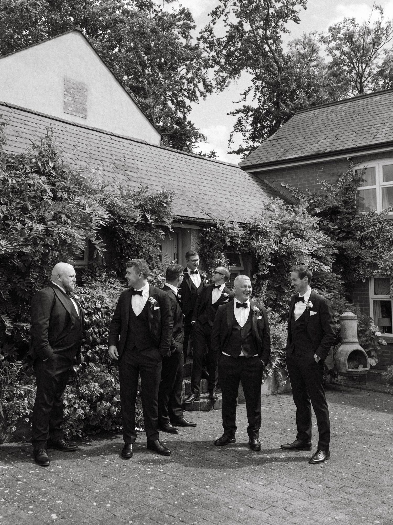 Groomsmen in black tuxedos gather outdoors for formal wedding photos.