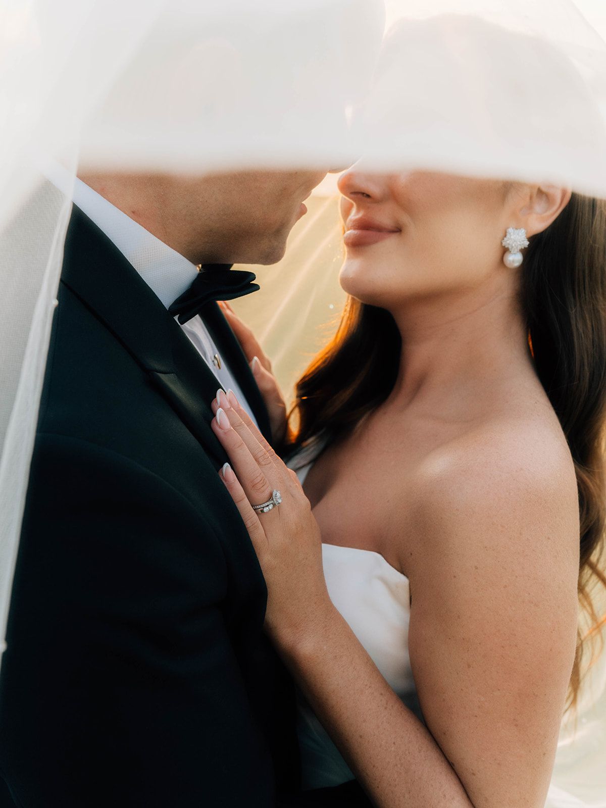 Close-up wedding photo showing a bride in white dress with pearl earrings embracing groom in black tuxedo.