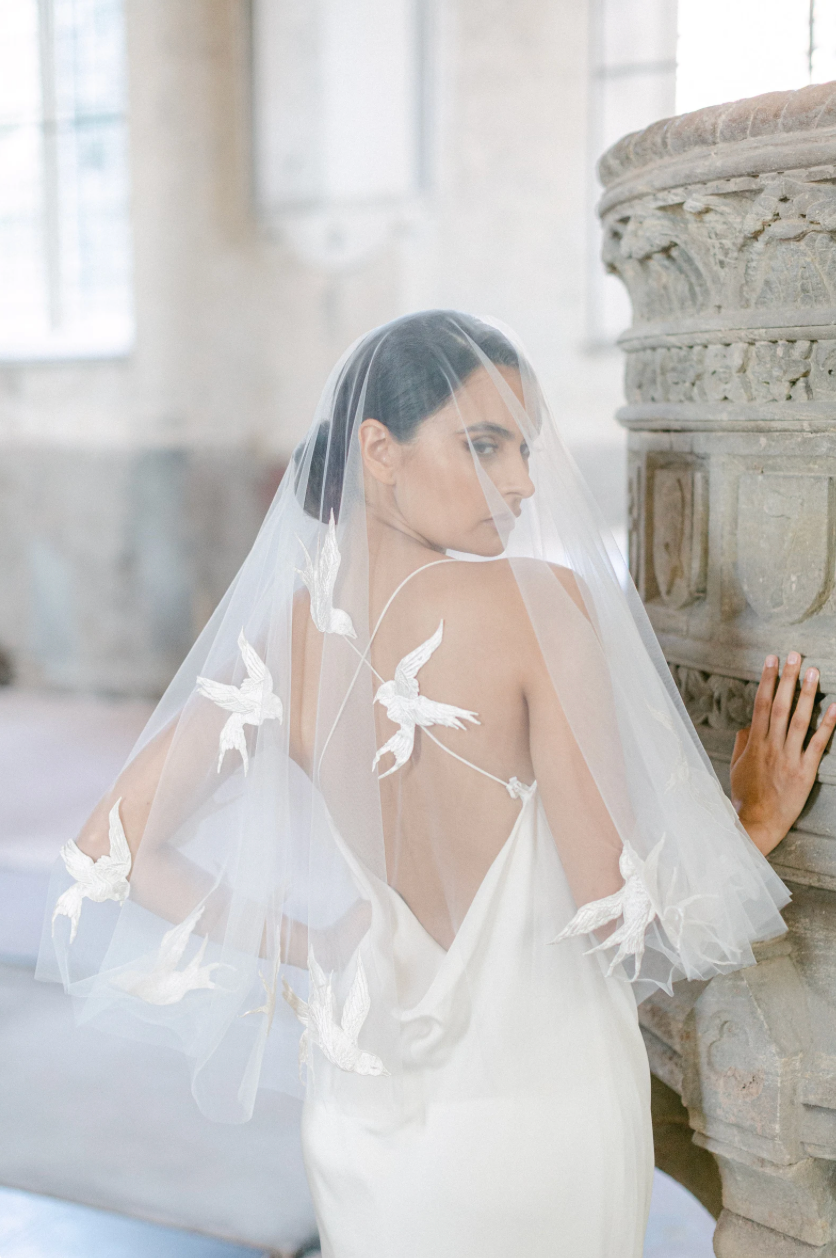 A bride in a flowing white veil with delicate bird appliques poses elegantly in a light-filled church interior.