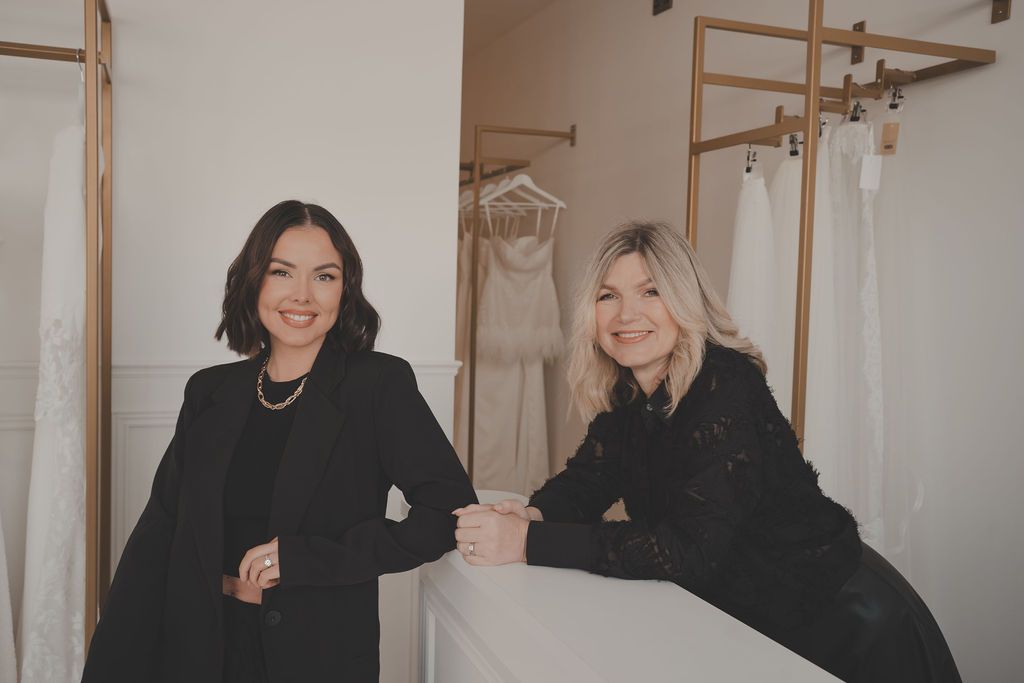 Two people in black outfits sit together in a room with a white dress hanging on display.