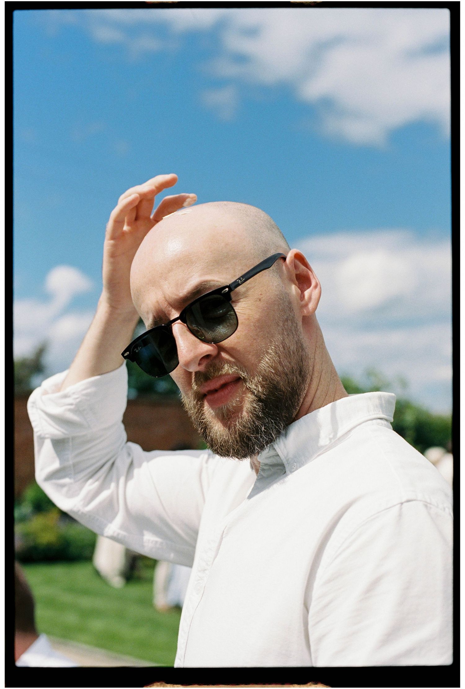 Bald man with beard wearing sunglasses and white shirt at outdoor summer wedding event.