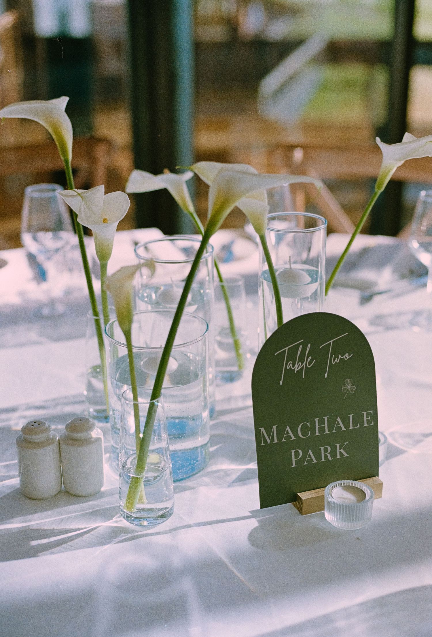 Elegant wedding table with white calla lilies in glass vases, a dark green table sign reading Table Two MacHale Park.