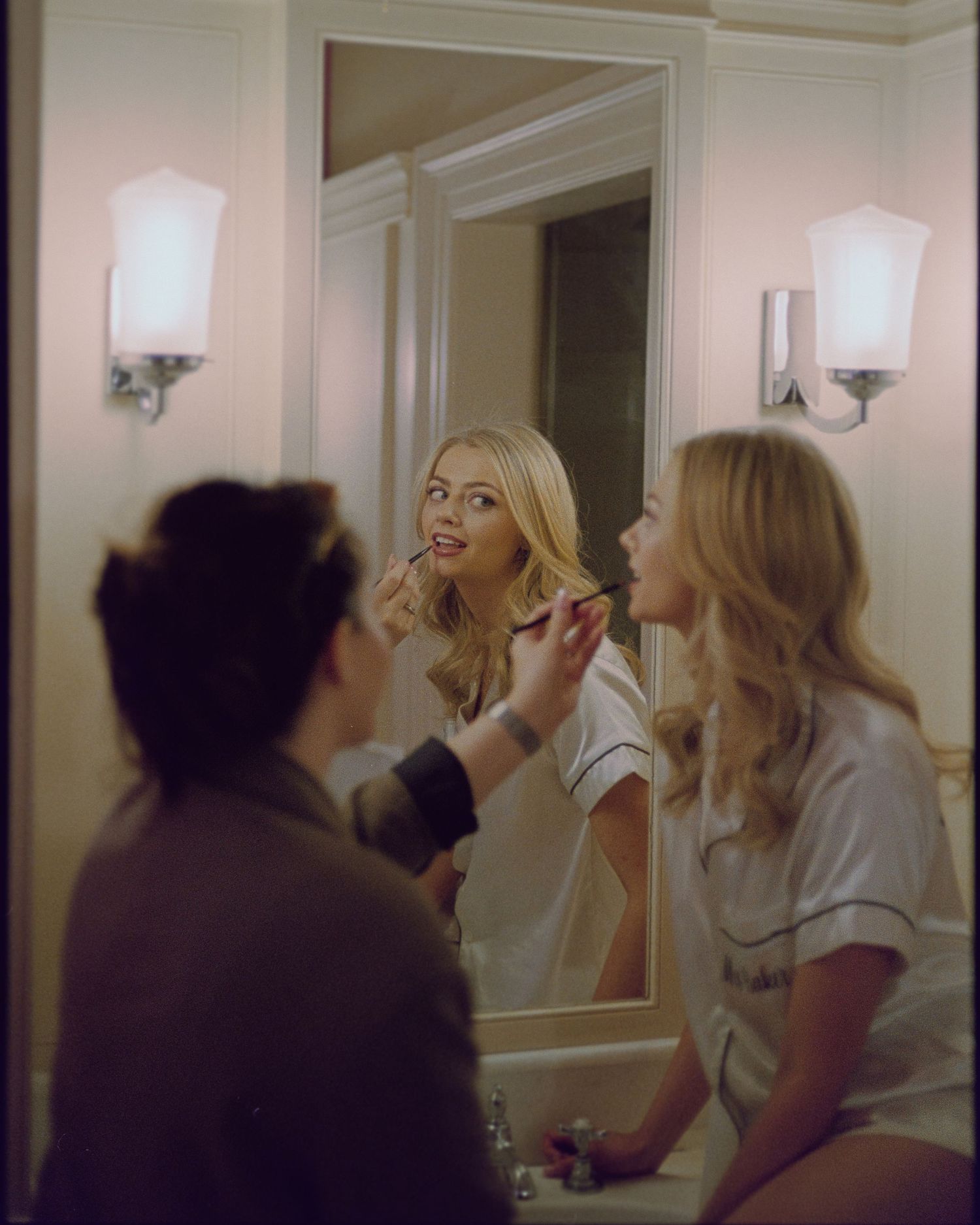 A makeup artist applies lipstick to a blonde woman in a white robe, reflected in a vanity mirror with elegant wall sconces.