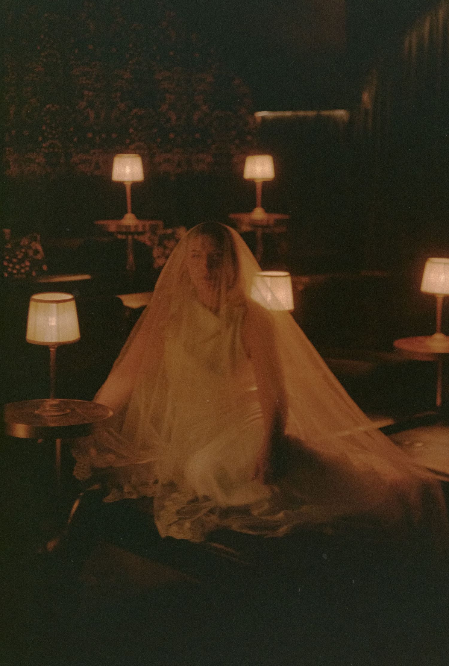 A figure draped in a flowing white veil sits in a dimly lit room surrounded by glowing table lamps.
