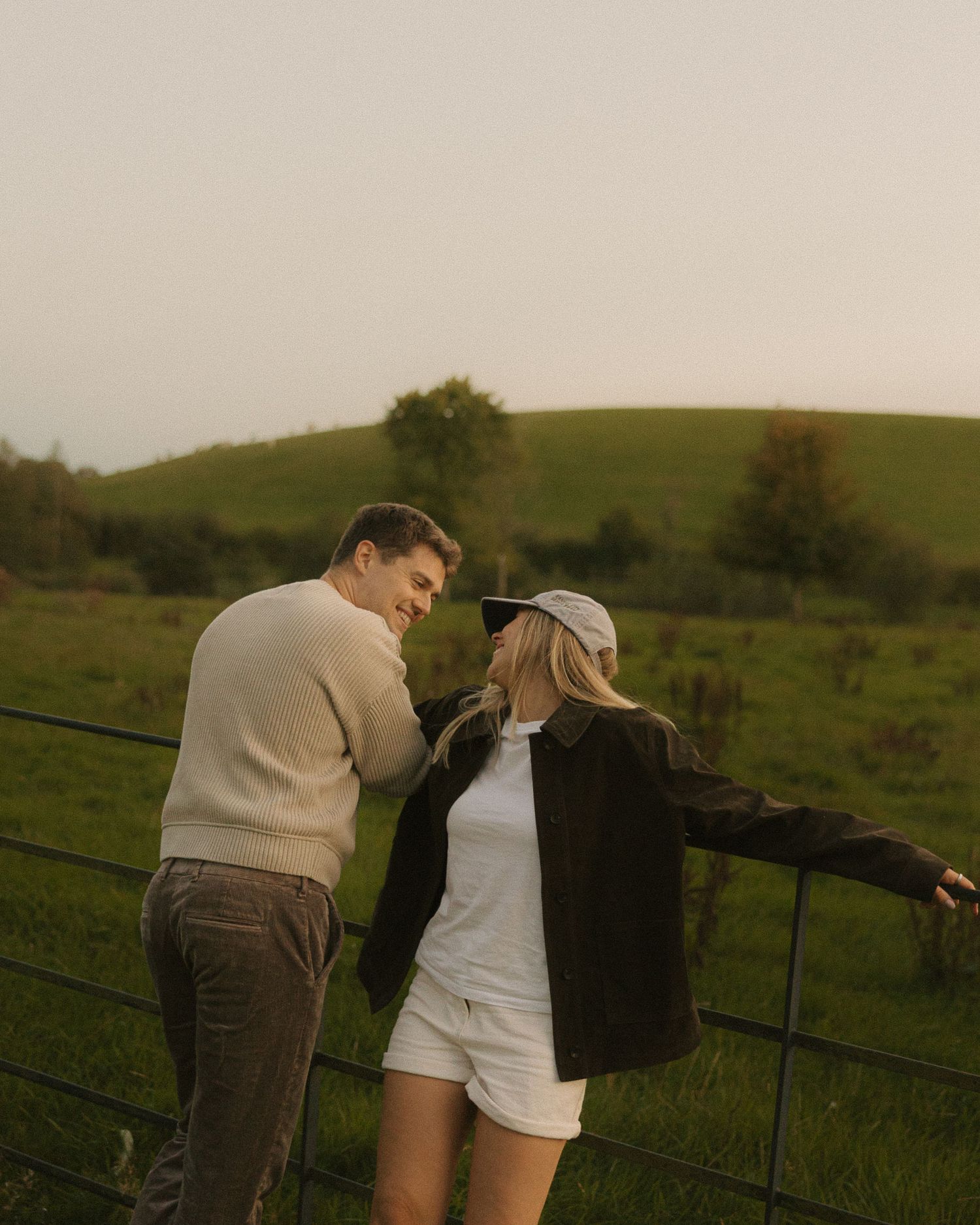Couple smiling at each other by a farm fence with lush green hills in the background at golden hour.