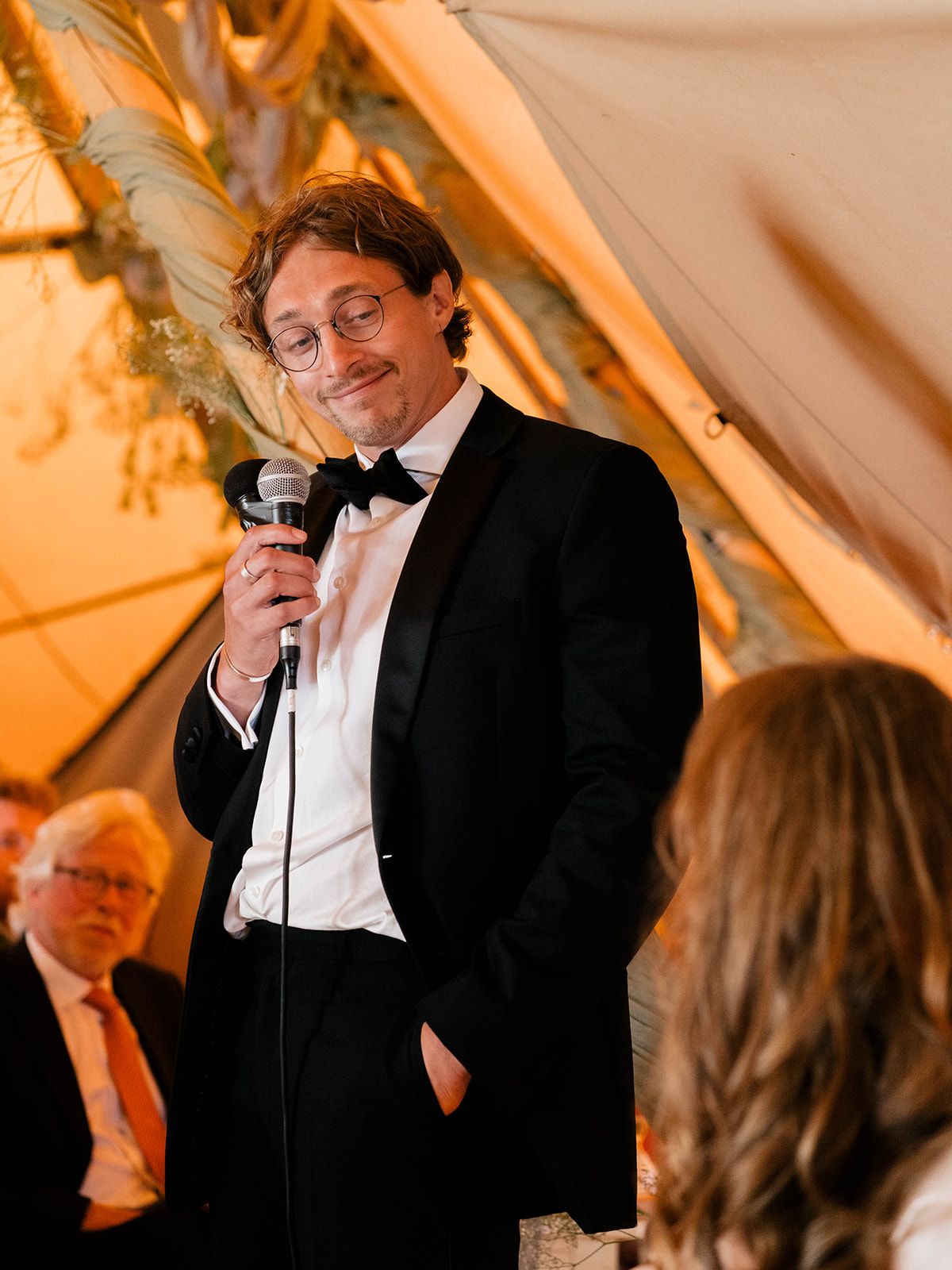 Groomsman in tuxedo gives heartfelt speech at wedding reception under tent lighting.
