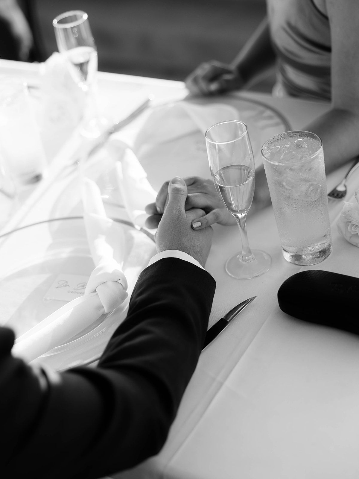 Two people holding hands across a white tablecloth with champagne glasses and beverages in black and white.
