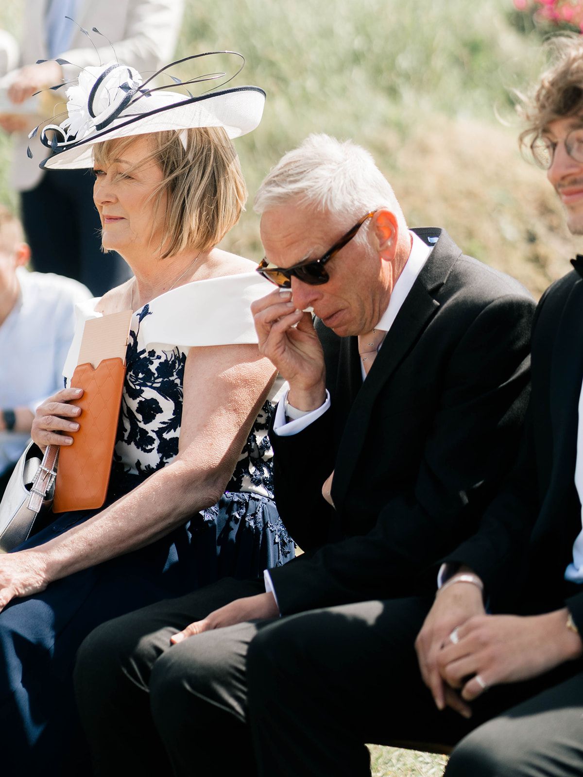 Wedding guests in formal attire sit outdoors during an emotional ceremony moment.