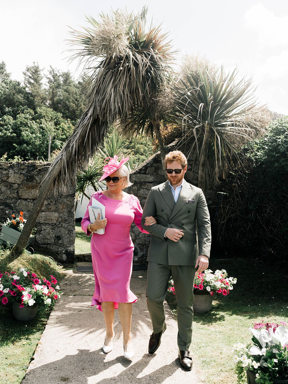 Two wedding guests walk along a garden path with palm trees and colorful flowers.
