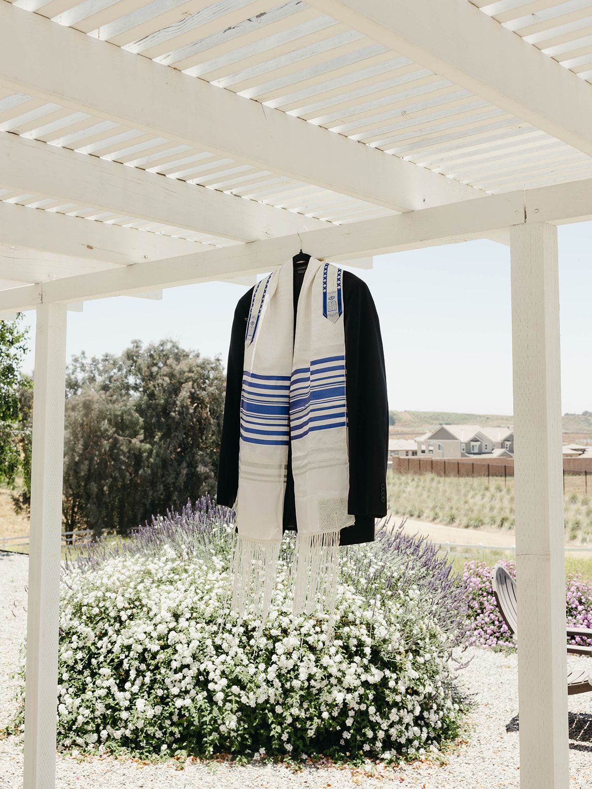 A white prayer shawl with blue stripes hangs from a pergola overlooking a lavender garden and desert landscape.