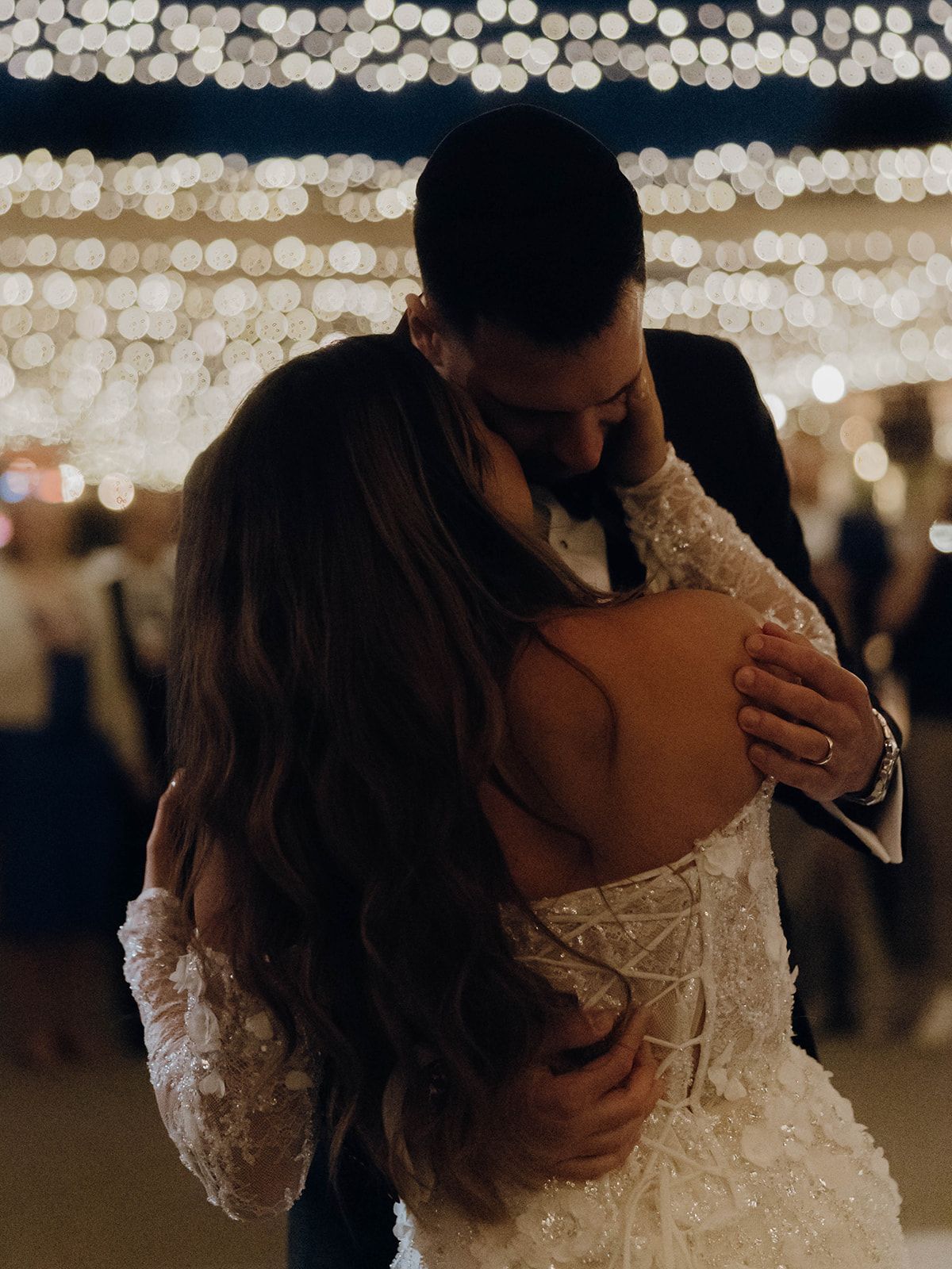 A romantic couple shares a tender embrace while dancing under twinkling lights at an evening celebration.