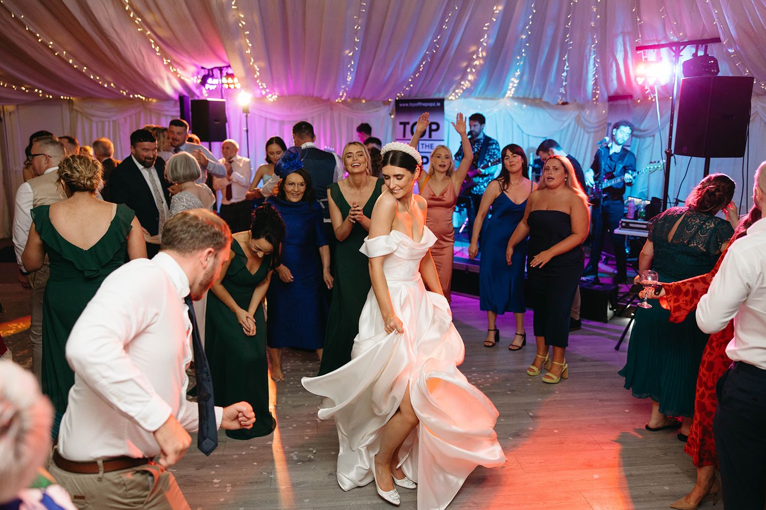 Wedding guests dance and celebrate on a lively reception dance floor illuminated by pink and blue lighting.
