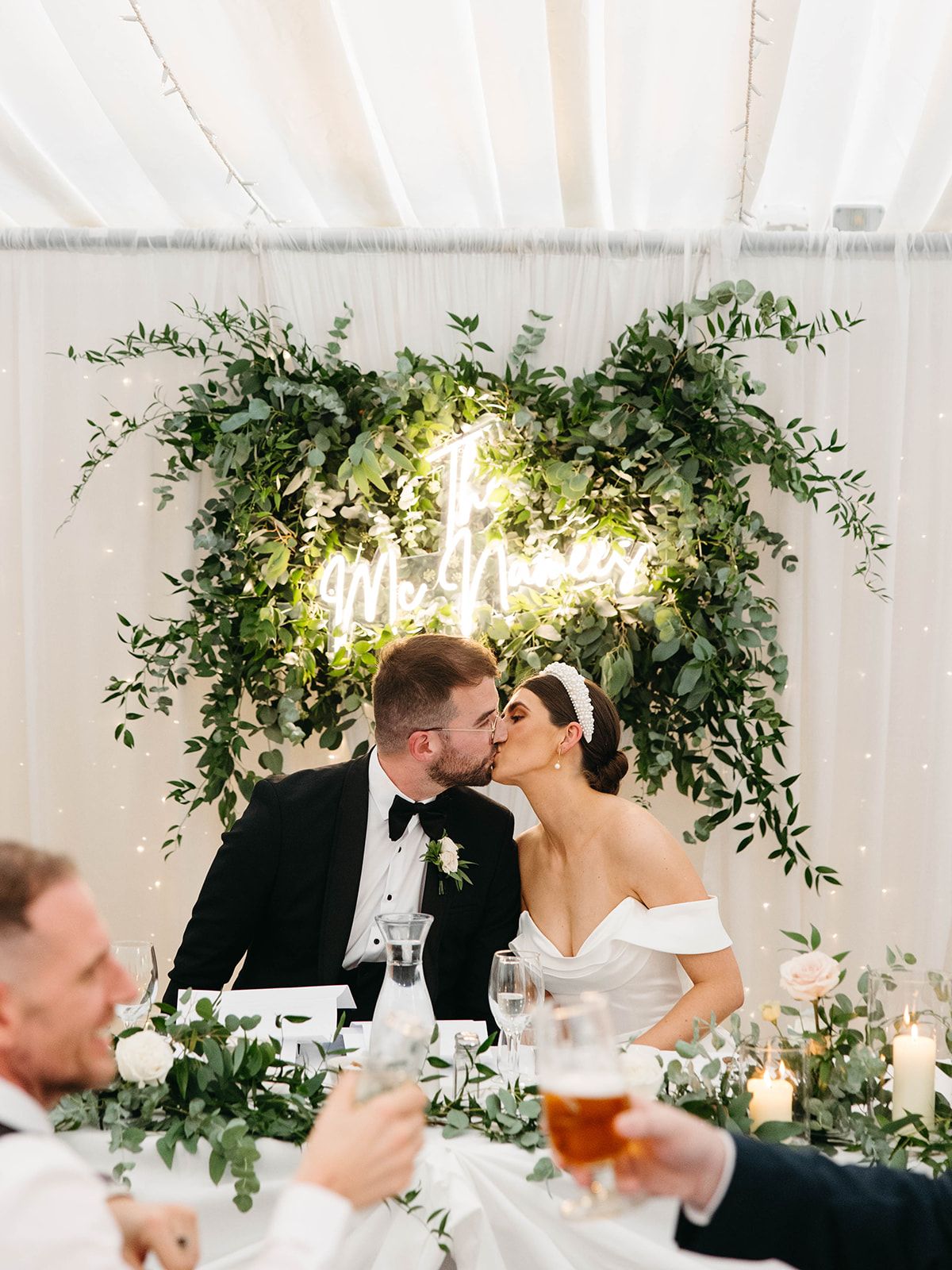 Romantic wedding reception scene with lit greenery arch and table decor as guests raise celebratory drinks.