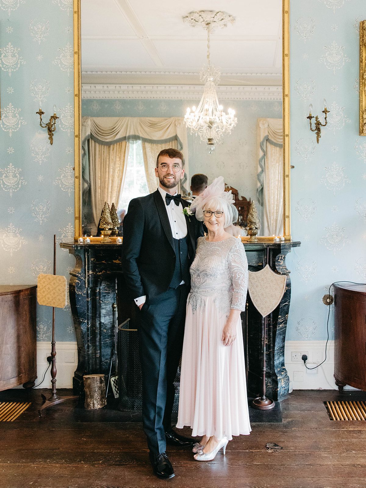 Elegant wedding photo in a vintage parlor room with ornate marble fireplace, crystal chandelier and soft blue walls.