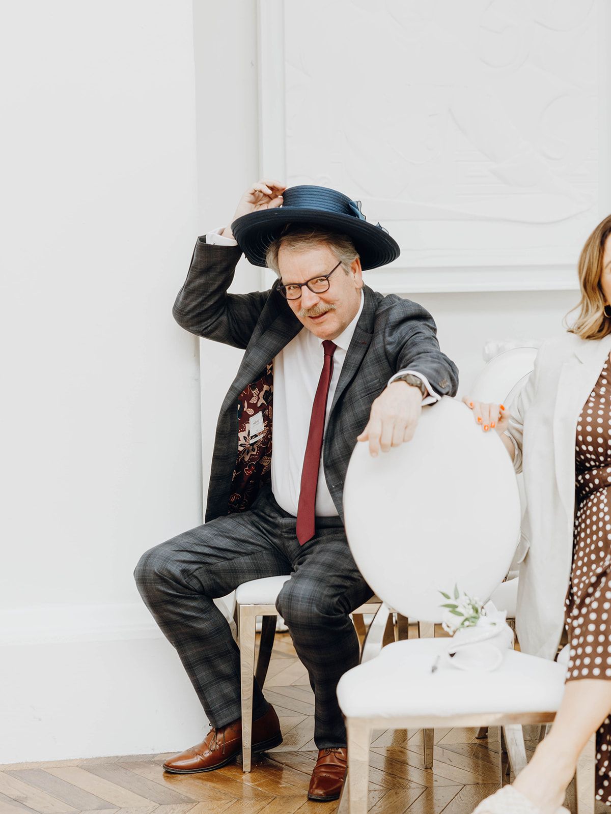 A person in a suit and hat sits on a white chair during what appears to be a wedding celebration or formal event.