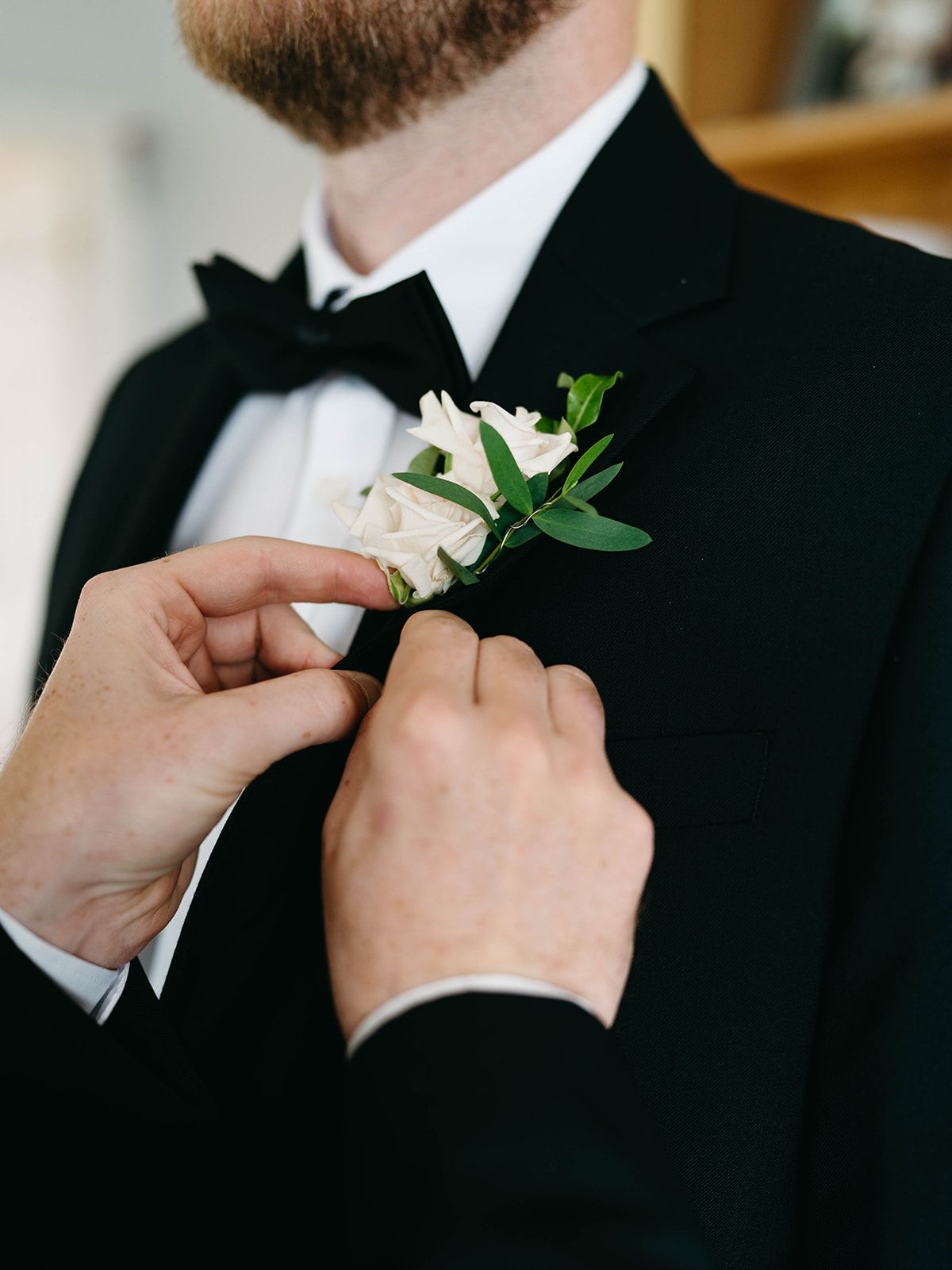 Someone pins a white corsage boutonniere with greenery onto a black tuxedo lapel during a formal event.