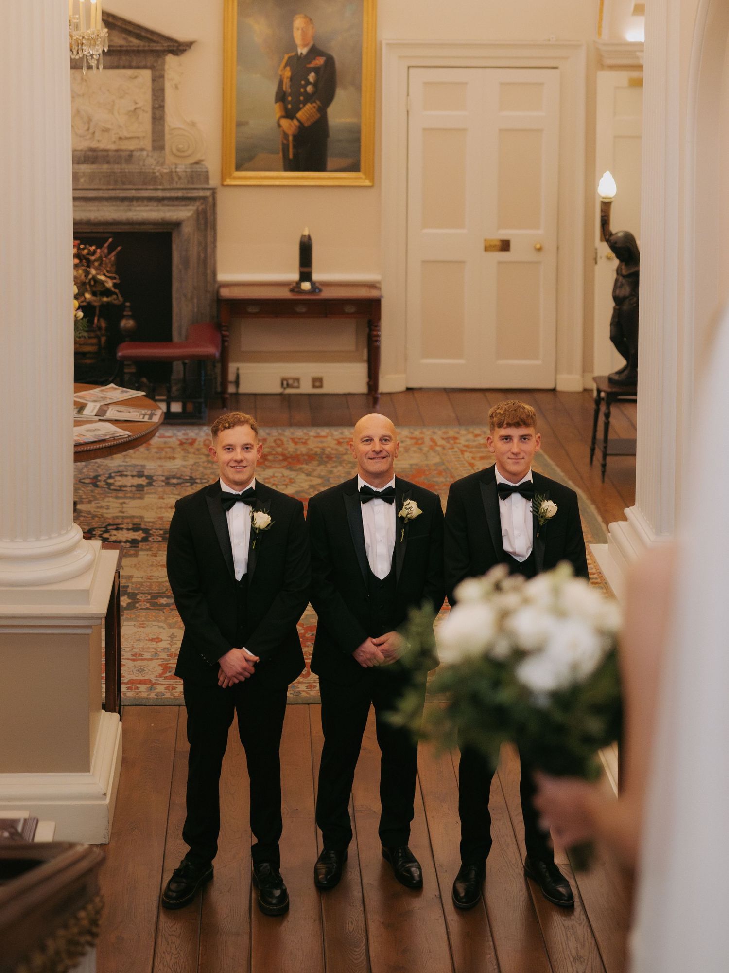 Groomsmen in black tuxedos and bow ties stand together in an elegant wedding venue with white walls and lighting.