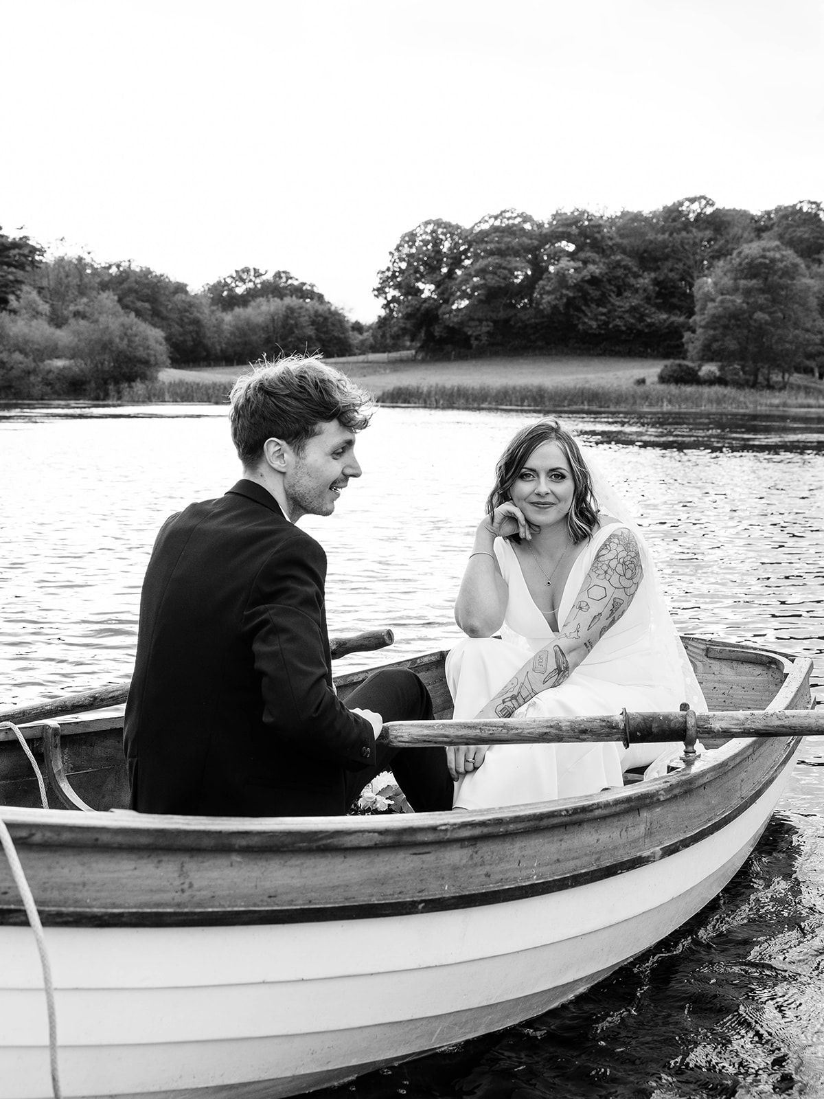 A couple share an intimate moment in a wooden rowboat on a tranquil lake surrounded by trees.