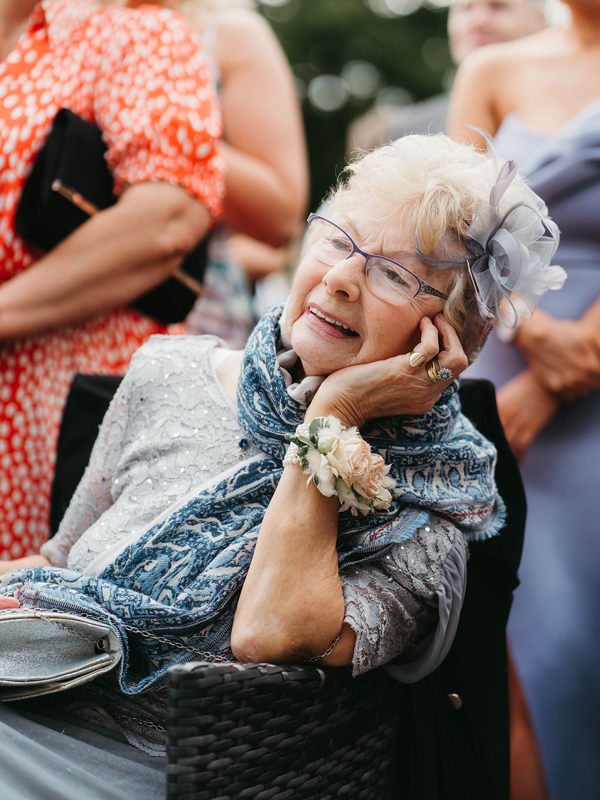 Guest in gray lace dress and blue scarf reacts with joy during outdoor event, wearing corsage and glasses.