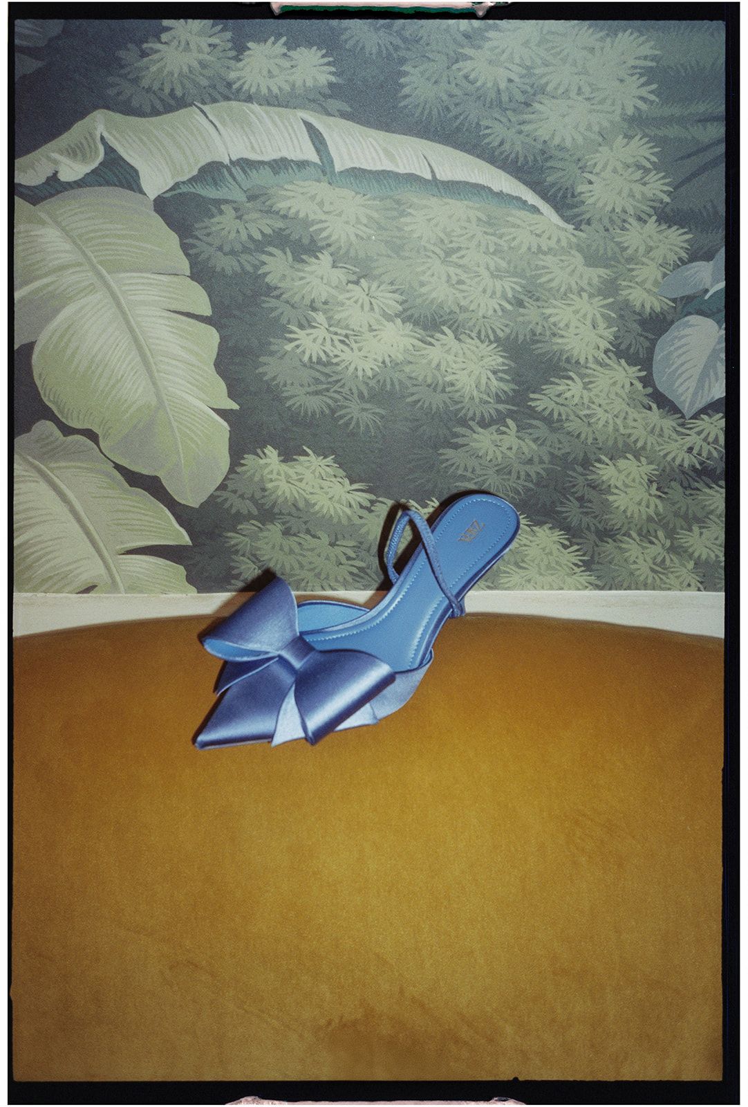A blue high-heeled shoe sits elegantly against a golden surface with a leafy backdrop creating an artistic composition.