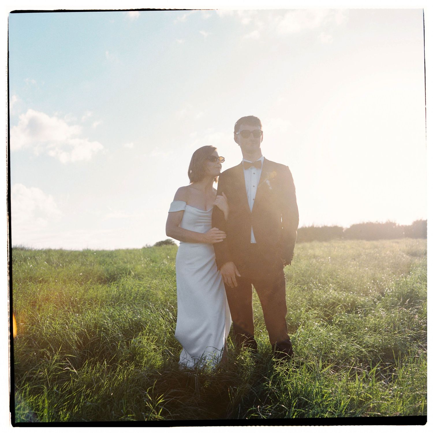 Bride in white gown and groom in dark suit standing together in a sunlit green field on their wedding day.